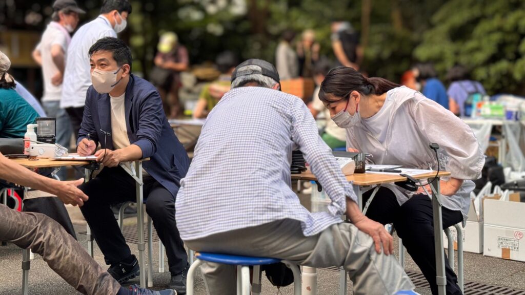 6月28日の炊き出し医療相談会の様子。
承知いたしました。画像の詳細な説明を含むALTテキストを生成します。これも前回の画像と同じ医療相談会であることを考慮に入れます。

やや高い位置から撮影された屋外の様子で、公園で開催されている医療相談会のブースが写っています。複数の人がテーブルを囲んで座っており、医療関係者と思われる人たちが相談に応じている様子です。中央手前には、白と水色のチェック柄のシャツを着た人が、背中を向けて青いスツールに座っています。その人の前には木目調の折りたたみテーブルがあり、書類やペンが置かれています。そのテーブルを挟んで、右側には灰色と白の市松模様のシャツとマスクを着用した、長い髪を後ろで束ねた人が、やや前かがみになってチェックシャツを着ている人と話しているか、書類に目を通しています。左側には、ベージュのマスクを着用し、ネイビーのジャケットを羽織った人が座っており、こちらも書類にペンを走らせています。画面左下には、茶色のズボンを履いた別の人物の足と手が写っており、テーブルの端に座っているようです。背景には、ぼやけていますが、他の人々や白いテント、さらに別のテーブルや段ボール箱のようなものが多数見えます。全体的に曇りの日で、明るい自然光が当たっています。